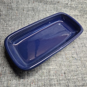 May include: A rectangular, navy blue ceramic dish with a glossy finish. The dish has a slightly raised, ridged border along the top edges. The interior is smooth and the corners are rounded. The dish is empty and sits on a grey, textured surface.