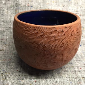 Può includere: Una ciotola in ceramica terracotta con interno smaltato blu. La ciotola ha un esterno testurizzato con un motivo geometrico.