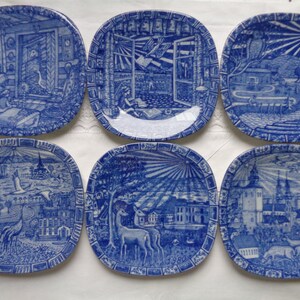 May include: Set of six blue and white ceramic plates with different scenes depicting a town, a house, a deer, a bird, and a sun. The plates are square and have a decorative border.