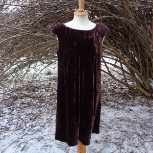 May include: A dark burgundy velvet dress with a round neckline and cap sleeves. The knee-length dress features vertical pleats. It is displayed on a mannequin. The setting is outdoors, with snow and bare branches.