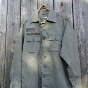 Puede incluir: Una camisa verde de manga larga con botones, un parche en el pecho izquierdo y dos bolsillos grandes. La camisa tiene un cuello abotonado y un ajuste holgado.