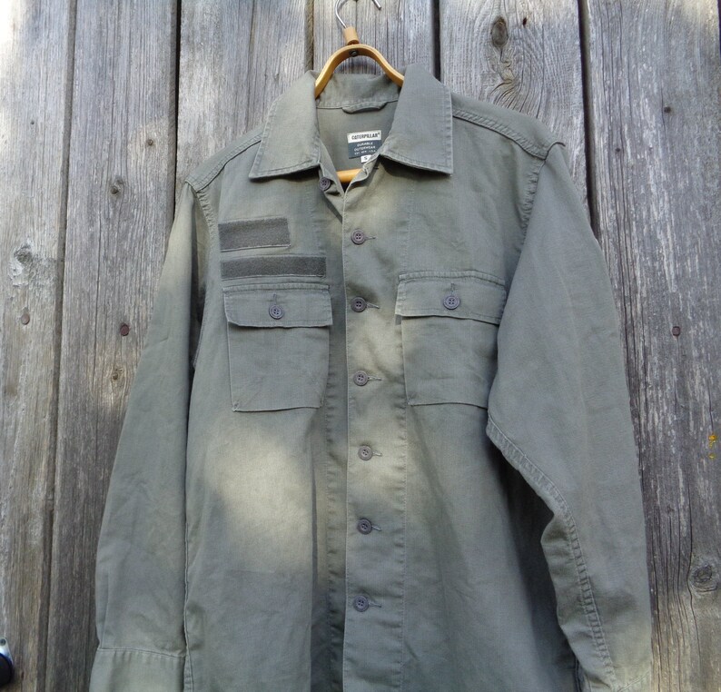 Puede incluir: Una camisa verde de manga larga con botones, un parche en el pecho izquierdo y dos bolsillos grandes. La camisa tiene un cuello abotonado y un ajuste holgado.