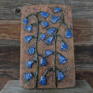 K&ouml;nnte beinhalten: Ein Wandbehang aus Keramik mit braunem Hintergrund und blauen Blumen. Die Blumen haben die Form von Glocken und sind vertikal angeordnet.