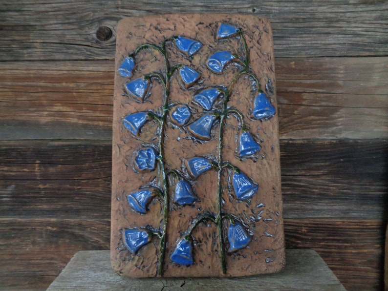 K&ouml;nnte beinhalten: Ein Wandbehang aus Keramik mit braunem Hintergrund und blauen Blumen. Die Blumen haben die Form von Glocken und sind vertikal angeordnet.