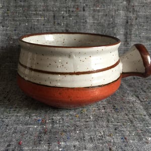 May include: A ceramic mug with a handle, featuring a white speckled body with two brown stripes and a burnt orange base. The rim and handle are also brown. The mug is designed for beverages.