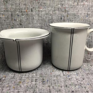 May include: Two white ceramic items with black trim and vertical black stripes. One is a mug with a handle, the other is a small bowl. Both have a simple, minimalist design.