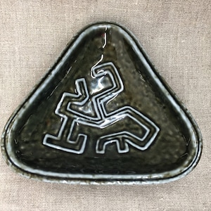 May include: A triangular ceramic dish with a dark green glaze and a white geometric design. The dish has a slightly raised rim.