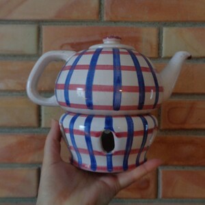 Op de afbeelding: Een witte theepot van keramiek met een blauw en roze gestreept ontwerp. De theepot heeft een ingebouwde verwarmer met een gat in het midden.