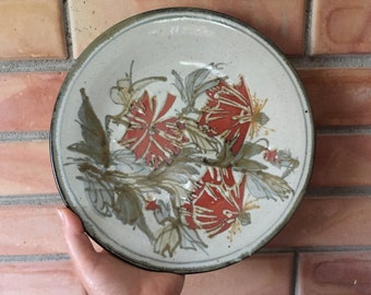 May include: A ceramic plate with a white background and a floral design. The flowers are red and brown and have a stylized look. The plate has a dark brown rim.