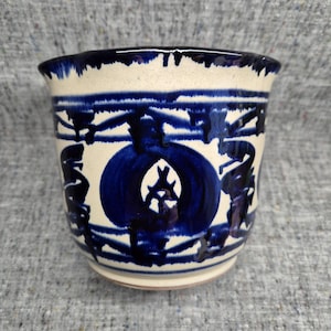 May include: A ceramic cup with a cream-colored base and blue painted designs. The cup features a large blue circle with a white center and other blue abstract shapes. The rim is also painted blue.
