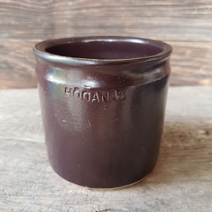 Op de afbeelding: Een donkerbruine keramische pot met het woord "HOGANAS" erop gegraveerd.