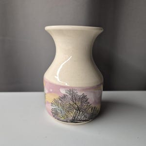 May include: A cream-colored ceramic vase with a flared top and a rounded body. The vase features a landscape design with a tree and a pink and yellow sky. The vase is suitable for displaying flowers or as a decorative item.