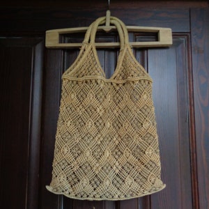 Op de afbeelding: Een beige gehäkelde touwtas die aan een houten hanger hangt tegen een donkerbruine houten deur.