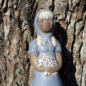 May include: A ceramic figurine of a girl wearing a blue dress and a white bonnet. She is holding a small white animal in her arms.