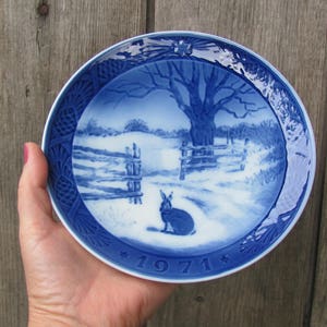 May include: Blue and white porcelain plate with a winter scene featuring a rabbit and a tree. The plate has a blue border with a white floral design and the year "1971" in the center.