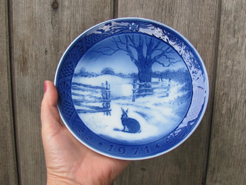 May include: Blue and white porcelain plate with a winter scene featuring a rabbit and a tree. The plate has a blue border with a white floral design and the year "1971" in the center.