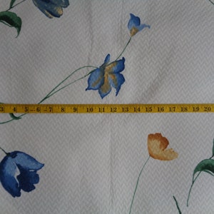 May include: White fabric with a repeating pattern of blue and orange flowers. A yellow measuring tape is placed across the fabric, showing measurements from 1 to 23 inches.