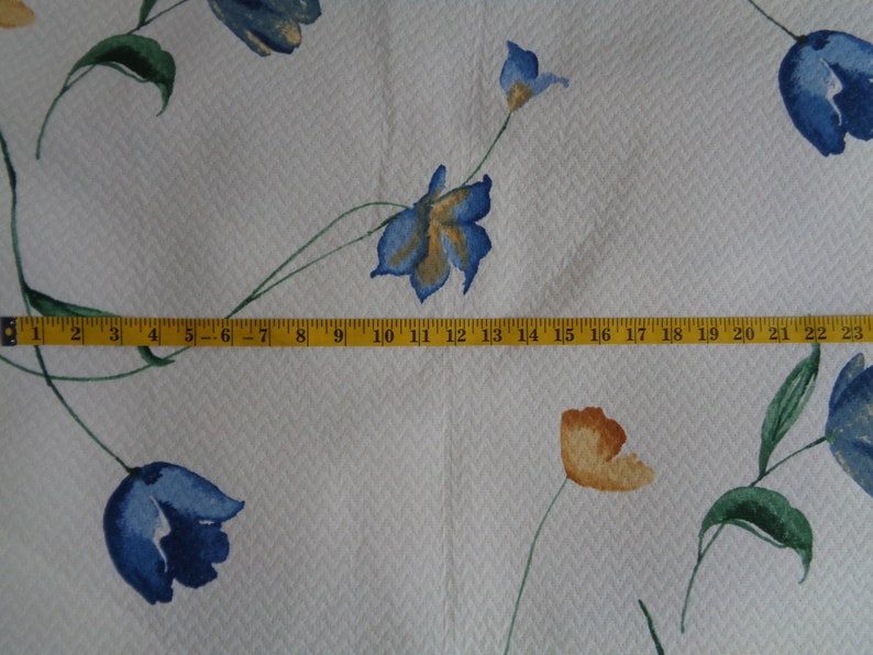 May include: White fabric with a repeating pattern of blue and orange flowers. A yellow measuring tape is placed across the fabric, showing measurements from 1 to 23 inches.
