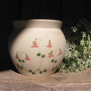 May include: A cream-colored ceramic pot with a rounded shape, adorned with a floral design featuring pink bell-shaped flowers and green stems. The pot sits on a wooden surface, with a cluster of white flowers to the right.