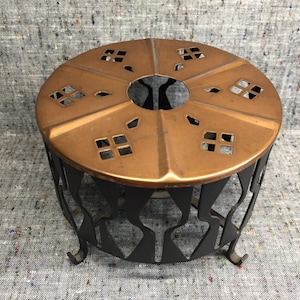 Op de afbeelding: Een koperkleurige metalen theewarmer met een decoratieve, uitgesneden bovenkant en een zwarte, sierlijke basis. De bovenkant heeft geometrische uitsparingen. De basis heeft een herhalend patroon van gebogen vormen.
