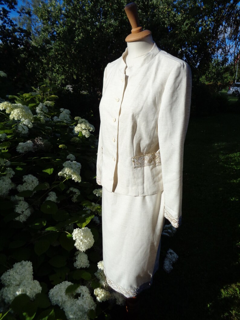 May include: A white linen suit with a mandarin collar and gold trim. The jacket has a button closure and the skirt has a straight silhouette.