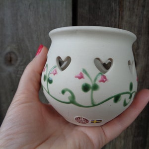 May include: A cream ceramic candle holder with heart-shaped cutouts and floral patterns. The holder is decorated with pink flowers and green vines. The Swedish flag is visible on the base.