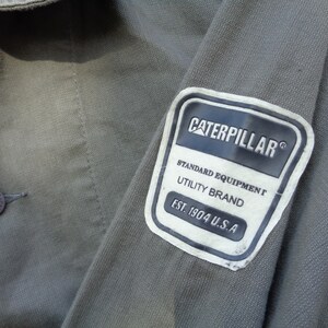 Puede incluir: Primer plano de una chaqueta de trabajo verde con un parche blanco que dice "Caterpillar&reg; Standard Equipment Utility Brand Est. 1904 U.S.A."