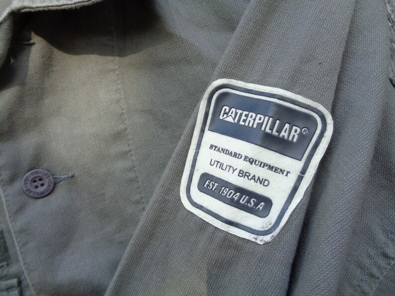 Puede incluir: Primer plano de una chaqueta de trabajo verde con un parche blanco que dice "Caterpillar&reg; Standard Equipment Utility Brand Est. 1904 U.S.A."