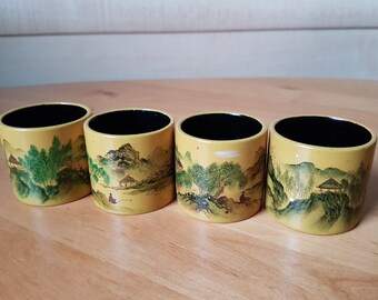 May include: Four small yellow cups with black interiors and hand-painted Asian landscape scenes. The cups are lined up in a row on a wooden surface.