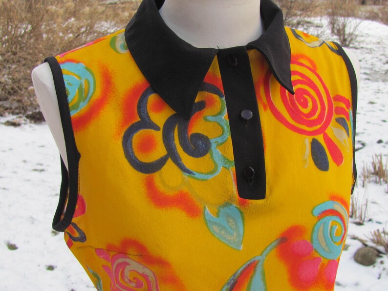 K&ouml;nnte beinhalten: Eine &auml;rmellose gelbe Bluse mit schwarzem Kragen und Kn&ouml;pfen. Die Bluse hat einen bunten abstrakten Druck mit Wirbeln und Blumen.