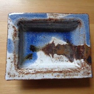 Op de afbeelding: Een rechthoekige keramische schaal met een blauwe, witte en bruine glazuur. De schaal heeft een verhoogde rand en een licht getextureerd oppervlak.