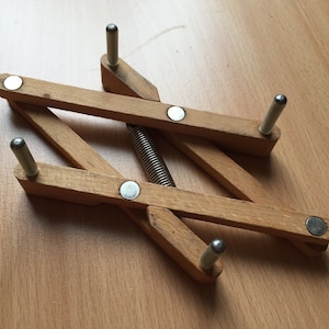 Op de afbeelding: Een houten vouwstandaard met een veermechanisme en metalen pinnen. De standaard is gemaakt van lichtbruin hout en heeft een natuurlijke afwerking.