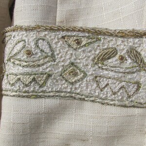 May include: A white fabric garment with a decorative trim of gold and silver embroidery. The embroidery features a repeating pattern of stylized shapes and geometric designs.