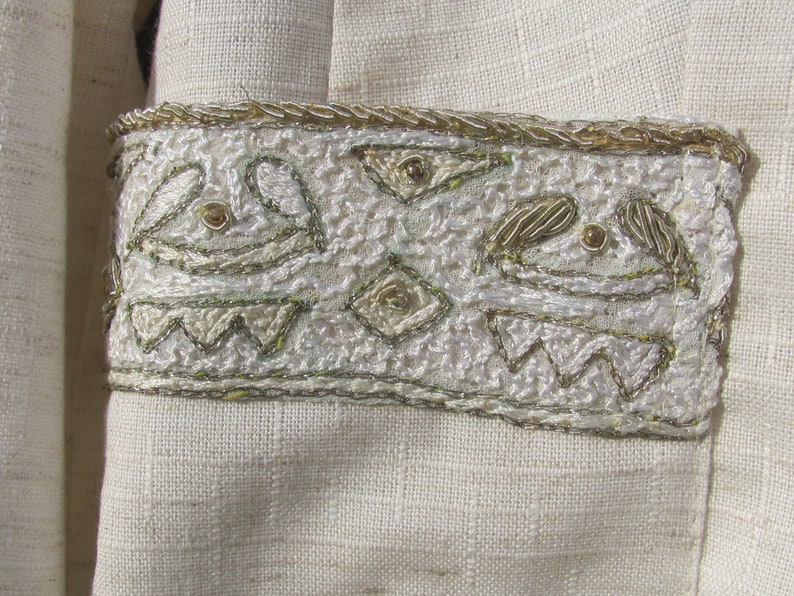 May include: A white fabric garment with a decorative trim of gold and silver embroidery. The embroidery features a repeating pattern of stylized shapes and geometric designs.