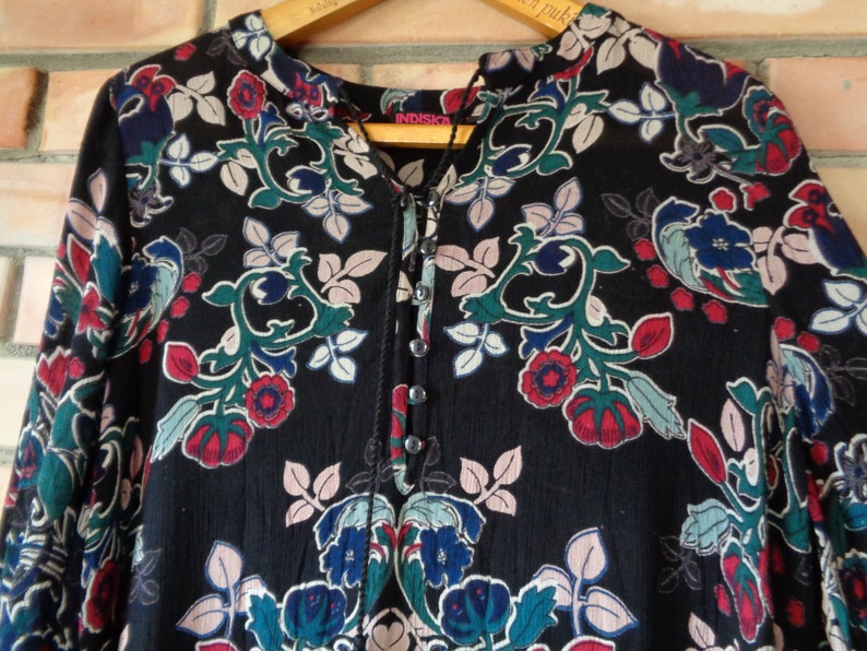 May include: Black long-sleeve shirt with a floral pattern in red, blue, and green. The shirt has a mandarin collar and a button-down placket. The brand name "INDISKA" is visible on the wooden hanger.