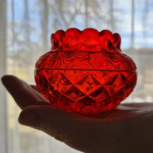 Op de afbeelding: Een helderrode glazen pot met een deksel, een geschulpte rand en een diamantgeslepen patroon. De pot heeft bloemreliëfdetails. De pot wordt in een hand gehouden, met een vage achtergrond.