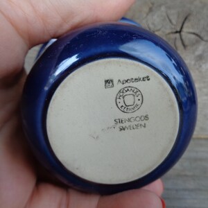 May include: A blue ceramic bowl with a white bottom. The bottom has the text "Apoteket", a logo with the text "Hogan&auml;s Keramik", and the text "Stengods Sweden".