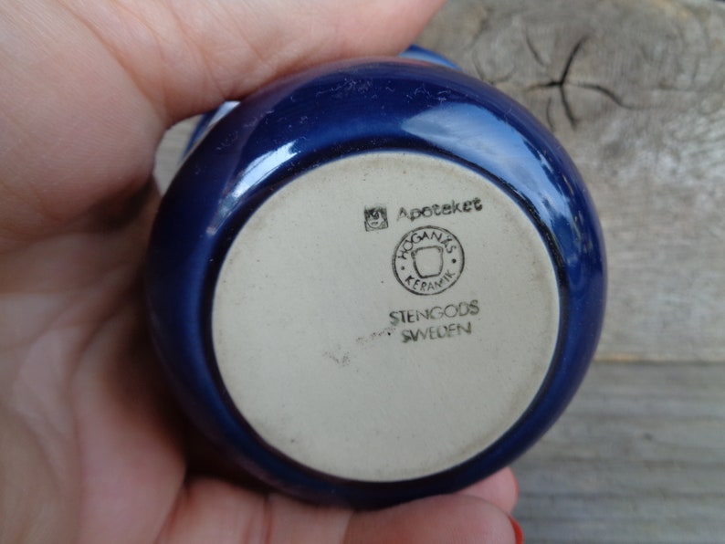 May include: A blue ceramic bowl with a white bottom. The bottom has the text "Apoteket", a logo with the text "Hogan&auml;s Keramik", and the text "Stengods Sweden".