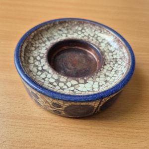 May include: A round ceramic candle holder with a blue rim and a textured, off-white and brown surface. The centre is a deep brown colour. The sides of the holder have a pattern of dark blue circles.