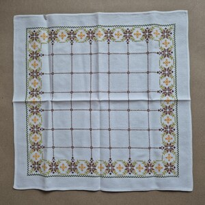 May include: White linen tablecloth with a cross-stitch design. The border features a repeating pattern of brown, yellow, and green floral motifs. The centre has a grid pattern of brown dotted lines.