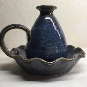 May include: A blue ceramic candle holder with a wavy base and a handle. The holder has a dark blue glaze with a slight sheen.