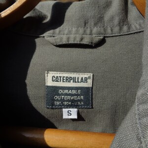 Puede incluir: Una camisa de trabajo verde de la marca Caterpillar con una etiqueta negra que dice "Durable Outerwear Est. 1924 - U.S.A." y una etiqueta de talla que dice "S".