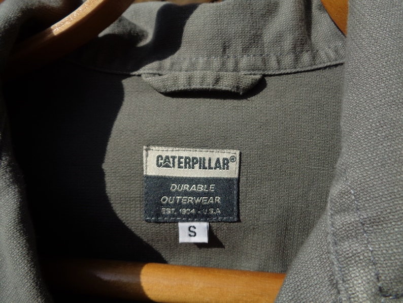 Puede incluir: Una camisa de trabajo verde de la marca Caterpillar con una etiqueta negra que dice "Durable Outerwear Est. 1924 - U.S.A." y una etiqueta de talla que dice "S".