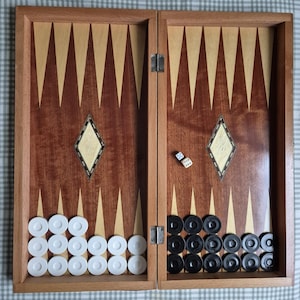 May include: A wooden backgammon board featuring a hinged design with a natural wood frame. The playing surface displays alternating dark and light wood triangular points, two diamond-shaped inlays, white checkers on the left, black checkers on the right, and two six-sided dice.