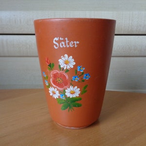 Könnte beinhalten: Eine Terrakotta-Tasse mit einem floralen Design, das auf die Vorderseite gemalt ist. Die Blumen sind weiß, blau und orange. Die Tasse trägt den Schriftzug "Säter" in weiß auf der Vorderseite.