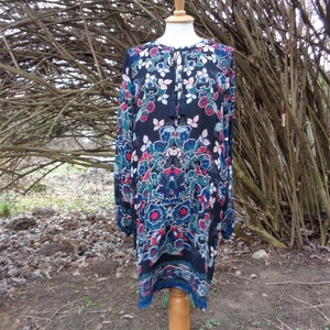 May include: A long-sleeved dress with a floral pattern in red, blue, and teal on a black background. The dress has a tie-neck detail and is displayed on a wooden dress form. The hemline is a contrasting blue.