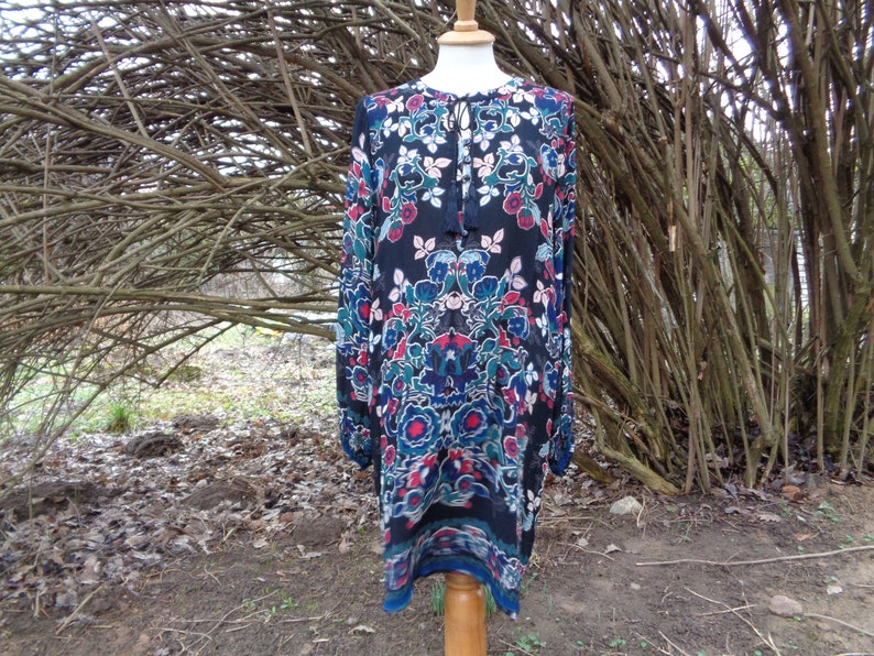 May include: A long-sleeved dress with a floral pattern in red, blue, and teal on a black background. The dress has a tie-neck detail and is displayed on a wooden dress form. The hemline is a contrasting blue.