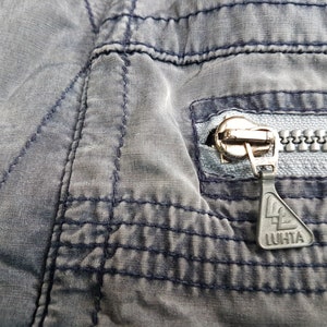 May include: Close-up of a zipper on a grey pair of trousers. The zipper pull has a black triangle with the word "LUHTA" printed on it.