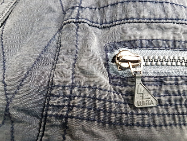 May include: Close-up of a zipper on a grey pair of trousers. The zipper pull has a black triangle with the word "LUHTA" printed on it.