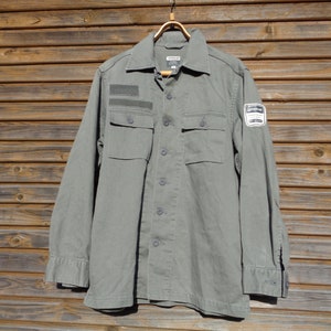 Puede incluir: Una camisa verde de manga larga con dos bolsillos y un parche en la manga derecha. El parche tiene el texto "WORKWEAR".